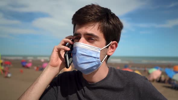 Young Man Answering A Phone Call On Beach While Wearing A Facemask Due To Pandemic. - close up