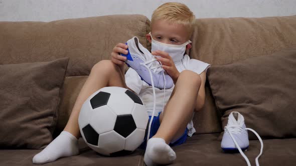 A Small Sad Boy in a Medical Mask Sits on a Sofa with a Soccer Ball and Cleats