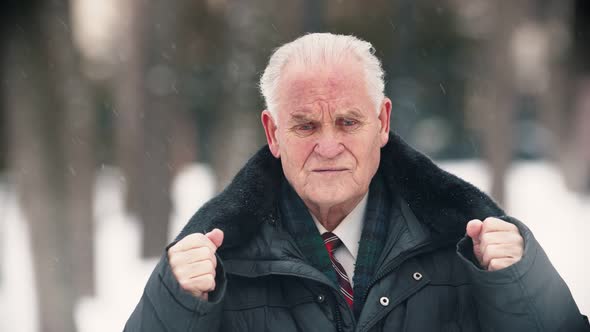 An Old Man Standing Outdoors While Snowfall - Putting His Hands Up and Touching His Head