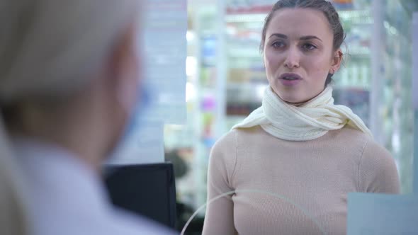 Young Sad Woman with Sore Throat and Migraine Talking to Druggist in Pharmacy Indoors