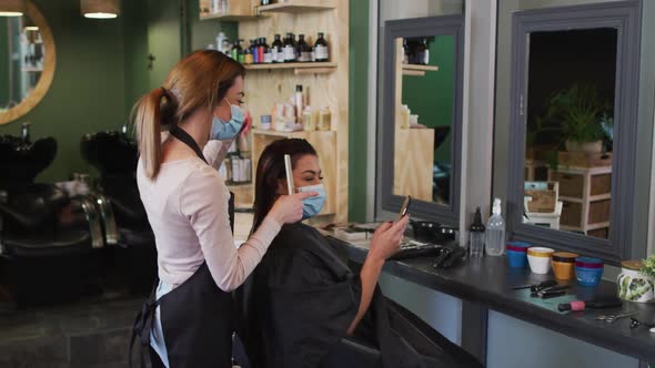 Female hairdresser and female customer wearing face masks looking at smartphone at hair salon