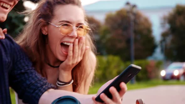 Happy Smiling Teenage Friends Laughing Outside at Something in Smartphone or Mobile Phone