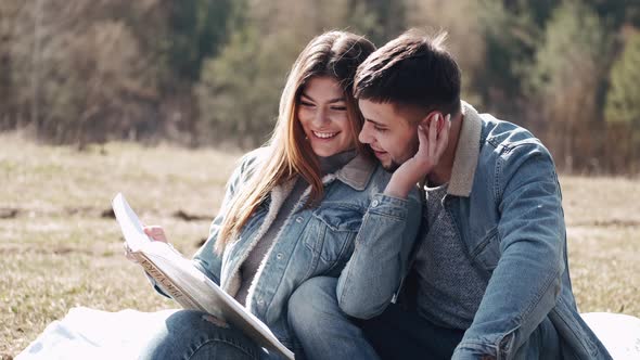 Attractive Young Man Is Hugging His Girlfriend Reading a Book in the Field