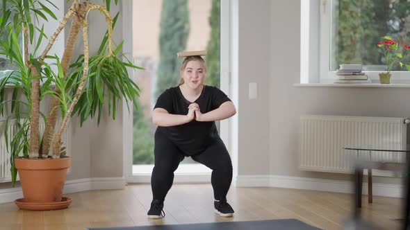 Wide Shot of Satisfied Smiling Plussize Woman in Squat Position with Book on Head Looking at Camera