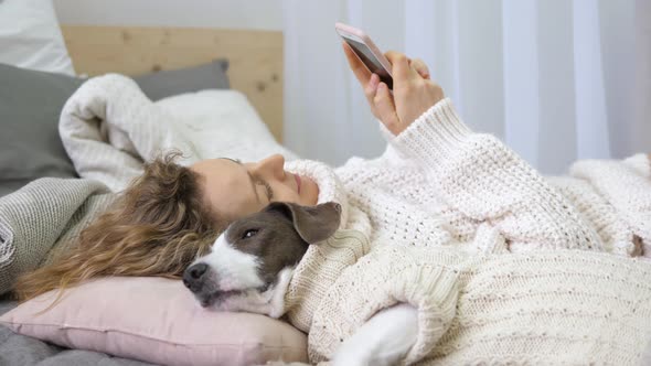 Girl Relaxing At Home With Her Pet Dog Using Mobile Phone In Comfy Cozy Clothes.