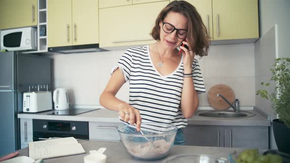 A Young Cheerful Woman Mixing Ingredients in a Bowl While Talking Her Phone