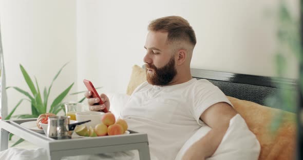 Side View of Bearded Guy Typing Message on His Smartphone While Sitting with Food Tray in Morning