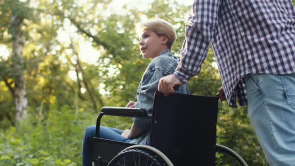 Young People with Disabilities in Wheelchair on Walk in Park with Her Loving Soulmate Side View