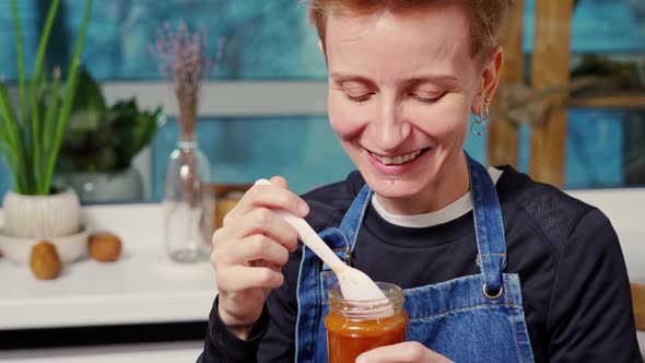a Cheerful Woman with Short Red Hair Eats Liquid Homemade Salted Caramel From a Jar with a Spoon and