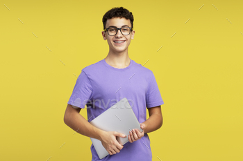 Smart school boy wearing eyeglasses holding laptop looking at camera isolated on yellow background