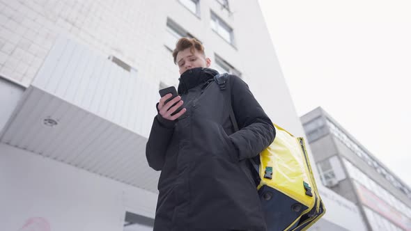Bottom Angle View of Focused Young Male Courier Standing Outdoors with Yellow Food Delivery Bag