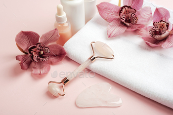 Facial cosmetic roller and gua sha scraper with pink orchid flowers on a white towel. Home spa