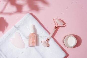 Facial cosmetic roller, gua sha scraper, serum bottle and cream jar on a white towel on pink
