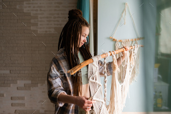 Young woman with dreadlocks weaves home decor from cotton thread macrame. 
