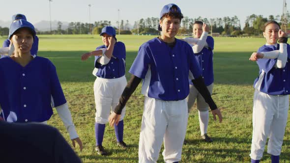 Diverse group of female baseball players and coach on pitch, warming up, stretching arms