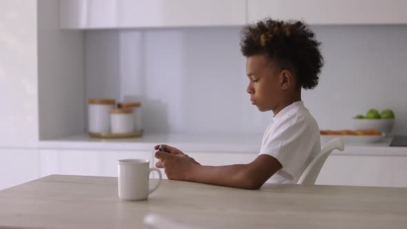 Handsome African American Boy is Using Smartphone at Table in Apartment Interior Spbi