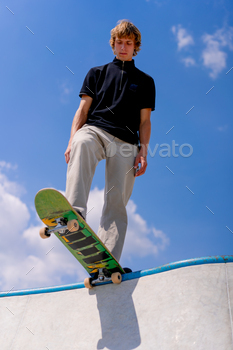 A young guy skater stands on the edge of a skate pool against a backdrop of sky and clouds