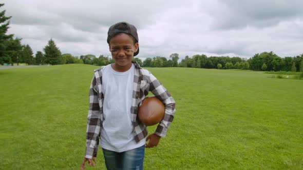 Happy African Little Boy with American Football Ball Walking on Pitch
