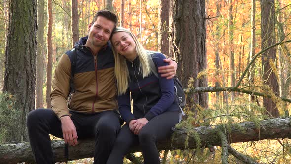 A Hiking Couple Sits on a Broken Tree in a Forest, Hugs and Smiles at the Camera