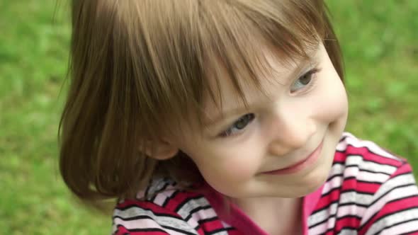 Close Up of Little Happy Girl in Summer Park Sits on Green Lawn Looks at Camera, Smiles and Laughs.