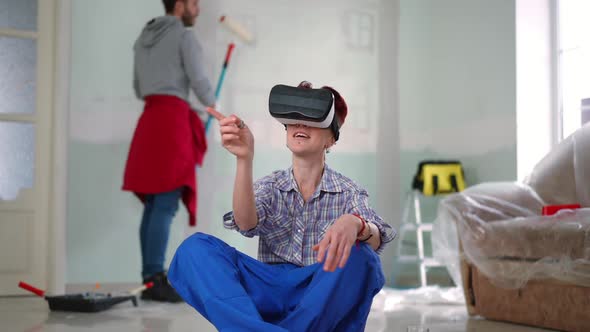 Smiling Woman in VR Headset Gesturing Examining Virtual Design As Man Painting Wall at Background