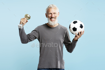 Attractive mature man, soccer player holding triumph cup and soccer ball, victory on blue background