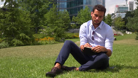 A Young Handsome Businessman Sits on Grass in a Park and Writes Notes - Office Buildings
