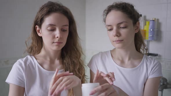 Funny Young Woman Touching Twin Sister Nose with Moisturizer Laughing As Siblings Applying Hand