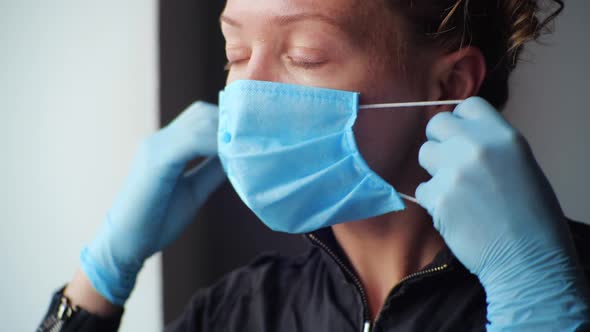 Young Millennial Woman Wearing Medical Protective Gloves Slowly Puts Disposable Face Shield Mask on