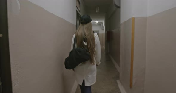 Back View of Young Slim Woman with Long Hair and Black Bag Passing Through Dim Corridor with Door