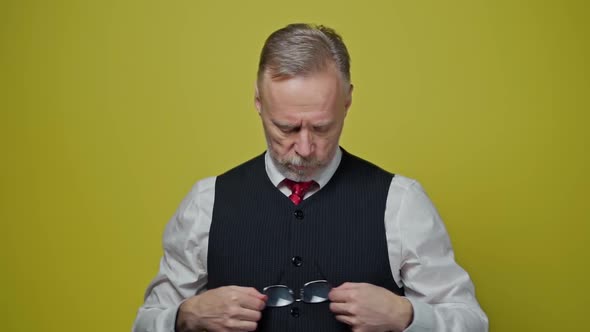 Senior man in elegant clothing holds glasses. Elderly man with grey beard wipes his glasses