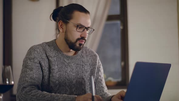 A Young Handsome Man in Glasses Works on a Laptop with a Glass of Wine