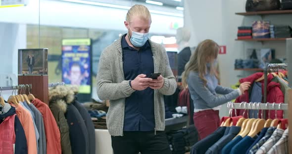 Confident Boyfriend in Covid Face Mask Messaging on Smartphone As Girlfriend Choosing Clothes in