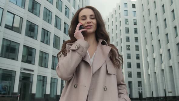 Cheerful young Caucasian woman standing in city street and texting on mobile phone with smile.