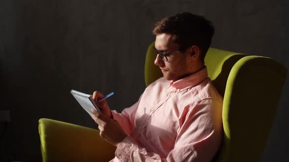 Tracking Shot of Thoughtful Young Man in Glasses Making Entries in Diary Sitting in Comfortable