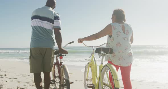 Smiling senior african american couple walking with bicycles on sunny beach