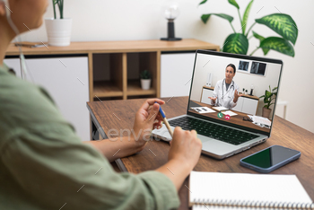 A person in a telemedicine session, writing notes while a doctor explains on a laptop