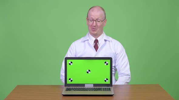 Happy Mature Bald Man Doctor Talking While Showing Laptop Against Wooden Table