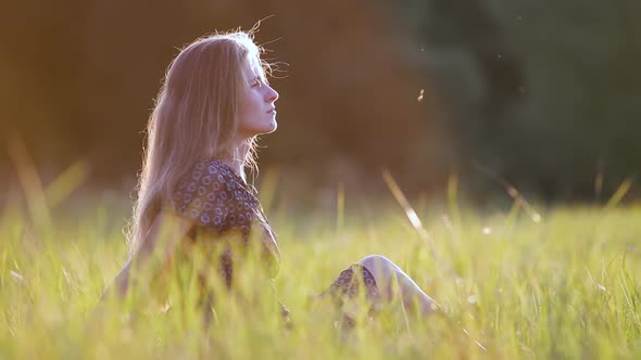 Young Woman with Long Hair Sitting Outdoors in Summer Field Grass Enjoying Nature at Sunset