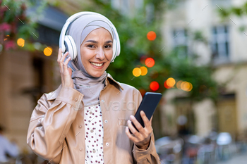 Smiling arabic woman in hijab listening audiobook in headset connected to mobile device while