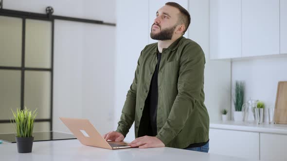 Portrait of Thoughtful Concentrated Caucasian Man Standing in Kitchen with Laptop