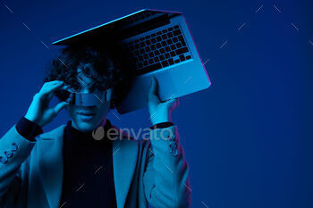 Portrait of stylish man hacker with laptop and futuristic glasses in blue light, cyber security