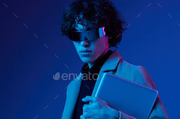 Portrait of a male hacker with a laptop and futuristic glasses in blue light, Blue Perennial color