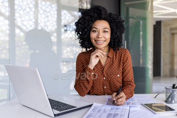Latino woman in polka dot blouse filling out tables by hand while working at technically equipped