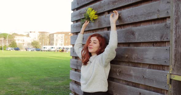 Young Girl with Bunch of Mimosa in Hand Smiles in a Park Near the Houses