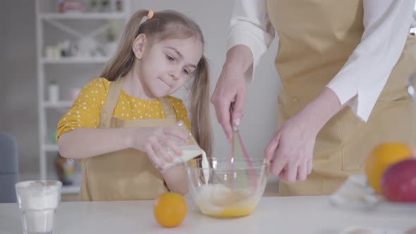 Cute Caucasian Brunette Girl Pouring Milk Into Bowl with Eggs. Unrecognizable Adult Woman Beating