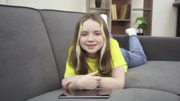 A Little Girl is Smiling While Lying on the Couch with a Tablet