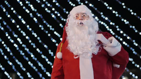 Santa Claus Showing Thumbs Up on the Background of Flashing Garlands Outdoors