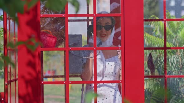 Beautiful Young Woman Happily Speaks on the Phone in an English Style Red Telephone Booth