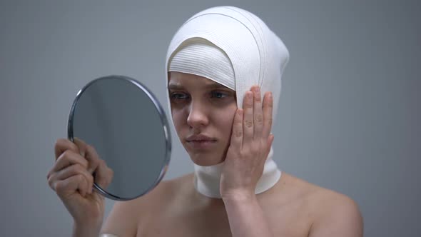 Female in Elastic Headwrap Looking in Mirror, Feeling Pain After Plastic Surgery